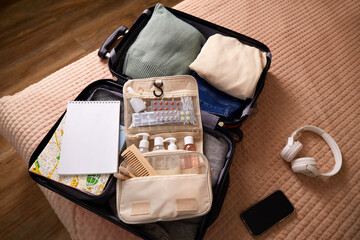 A suitcase is open on a bed, containing neatly arranged clothing, toiletries, and travel necessities. A notepad, headphones, and a smartphone are nearby, indicating preparation for a journey.