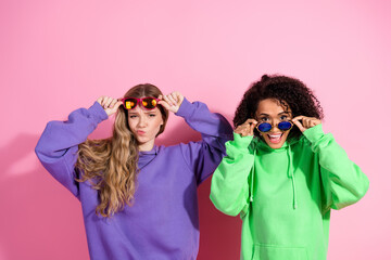 Two stylish girlfriends posing joyfully in colorful hoodies with sunglasses against a pink backdrop