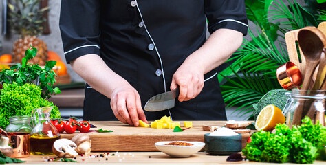 Chef In uniform cuts yellow potatoes into cubes. Cozy kitchen with wooden table, kitchenware, vegetables, herbs and ingredients for cooking. Healthy vegan food, culinary, recipes, food blogging
