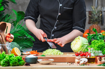 Chef brushes chopped cabbage off the knife. Cozy kitchen with wooden table, kitchenware, vegetables, herbs and ingredients for cooking. Healthy vegan eating, culinary, recipes, food blogging