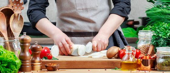 Home chef cuts white radish with santoke knife. Cozy kitchen with wooden table, kitchenware, vegetables, herbs and ingredients for cooking. Healthy vegan eating, culinary, recipes, blogging