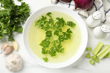 Tasty chicken bouillon in bowl and ingredients on white marble table, flat lay