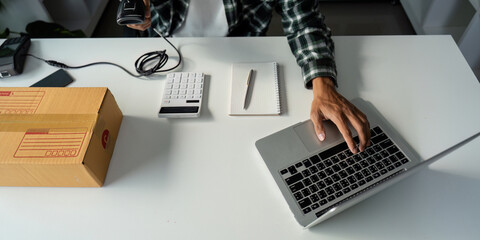 Person using laptop and calculator while managing online order fulfillment with barcode scanner and cardboard box.