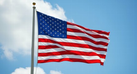 Fototapeta premium American flag waves against a bright blue sky with fluffy clouds.