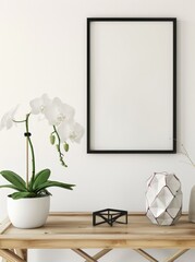 Fototapeta premium A white orchid plant in a white pot. a black frame. and a geometric white vase on a wooden table against a white wall