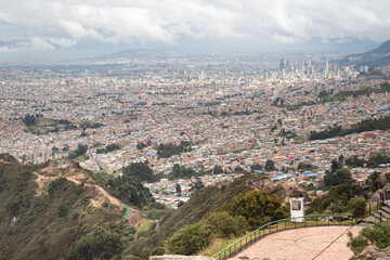 A panoramic aerial view of Bogotá, a sprawling city with mixed low-rise and high-rise buildings, green spaces, and a distant skyline under a cloudy sky
