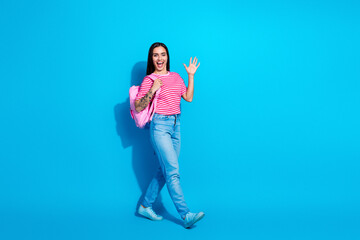 Smiling young woman waving with enthusiasm, wearing casual and stylish striped t-shirt and jeans, carrying a pink backpack
