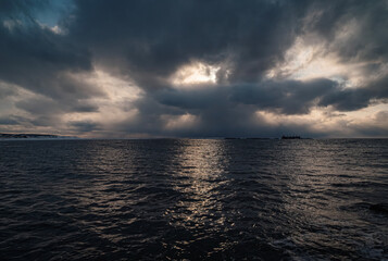 Dark clouds loom over the ocean during dusk, with rays of sunlight piercing through.