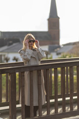 Woman in beige trench coat adjusts sunglasses on wooden bridge in peaceful town setting