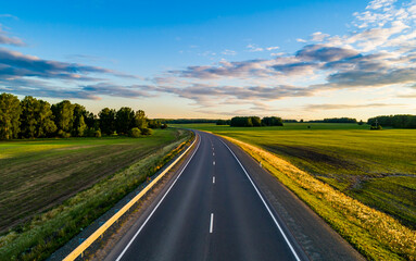 A winding road stretches into the distance, flanked by vibrant green fields and dense trees under a clear blue sky.