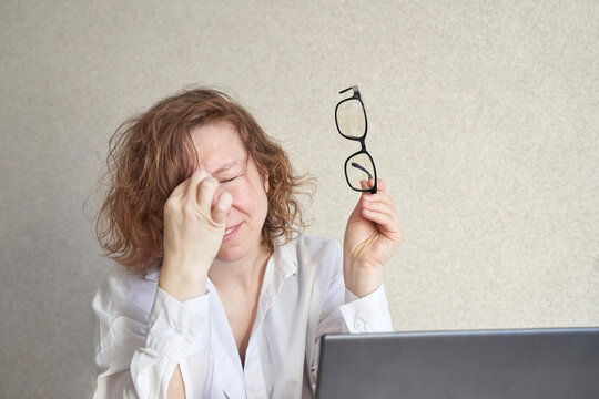 Caucasian female adult experiencing eye strain holding glasses near laptop while rubbing eyes due to screen fatigue.