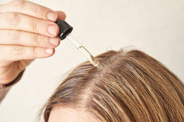 Female applying hair serum with dropper on scalp for hair care routine.