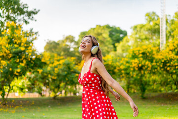 Obraz premium Young Woman in Retro Red Polka Dot Dress Wearing Headphones Listening and Enjoying Music in a Park Happily, Immersing with Nature