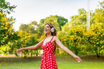 Young Woman in Retro Red Polka Dot Dress Wearing Headphones Listening and Enjoying Music in a Park...