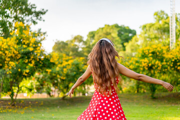 Young Woman in Retro Red Polka Dot Dress Wearing Headphones Listening and Enjoying Music in a Park...