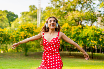 Young Woman in Retro Red Polka Dot Dress Wearing Headphones Listening and Enjoying Music in a Park...