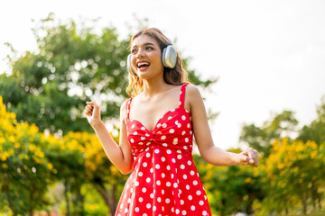 Young Woman in Retro Red Polka Dot Dress Wearing Headphones Listening and Enjoying Music in a Park...