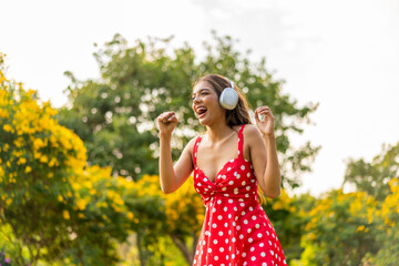 Young Woman in Retro Red Polka Dot Dress Wearing Headphones Listening and Enjoying Music in a Park...