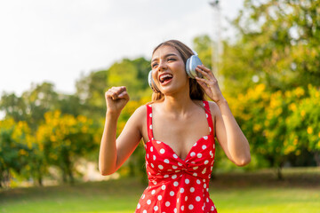Young Woman in Retro Red Polka Dot Dress Wearing Headphones Listening and Enjoying Music in a Park...