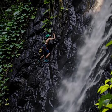 A mid-range shot captures a climber navigating a wet rock face near a cascading jungle waterfall, glistening water catching warm tropical light, mist wrapping around the scene.
