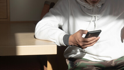 A person in a white hoodie sits at a table, holding a smartphone, with sunlight casting shadows around them.