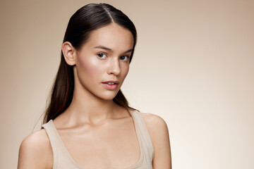 Fototapeta premium Portrait of a young woman with long hair wearing a light tank top against a soft beige background, conveying beauty and confidence