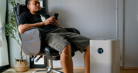 Pretty man sitting near air purifier and moisturizer appliance near sofa monitoring air quality in phone. 