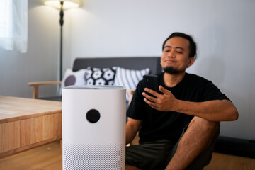 Pretty man sitting near air purifier and moisturizer appliance near sofa monitoring air quality in...