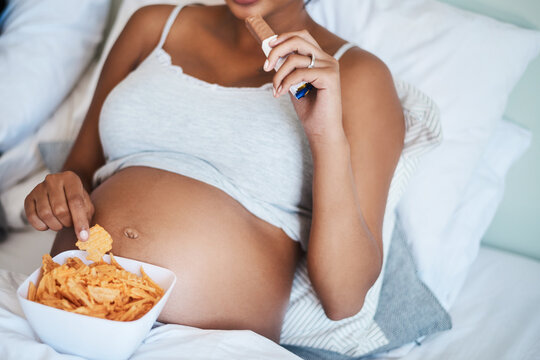 Pregnant, hands and woman with snack, home and craving for chocolate in bedroom, delicious and relax. Above, eating and person with chips on bed, stomach and future mom with sweet tooth in apartment - Powered by Adobe