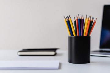 Fototapeta premium Workplace Stationery: A collection of colorful pencils sits neatly in a black holder, alongside a notebook and a partially visible laptop, on a clean workspace