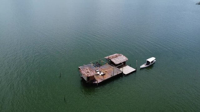 A floating bamboo hut with thatched roofs on Lake Tondano, Minahasa, Indonesia. A small boat is moored nearby, surrounded by calm, greenish water under a clear sky