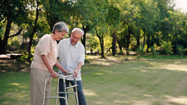 Asian senior husband take care to wife having knee pain while walking with walker stick in the park, Physical therapy in outdoor concept
