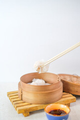 Steamed Hakau Dumpling Held By Chopsticks Over Bamboo Steamer With Chili Sauce Dipping Bowl On Wooden Mat For Authentic Asian Culinary Experience.