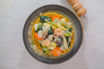 Ifumie Topped With Shrimp, Fresh Vegetables, And Thick Savory Gravy Served In Grey Bowl On Wooden Trivet With Pepper Grinder In Background.