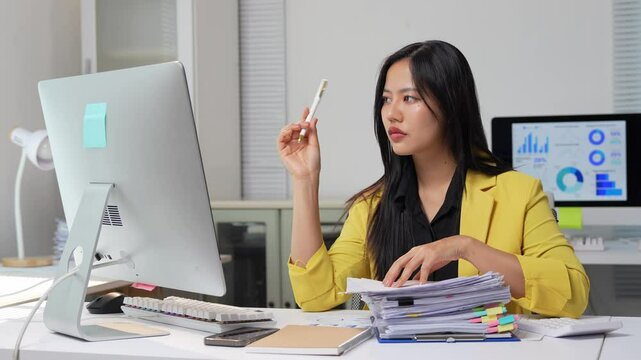 Young asian businesswoman wearing yellow jacket sits at desk in office, looking at computer screen and holding pen, with stack of documents nearby