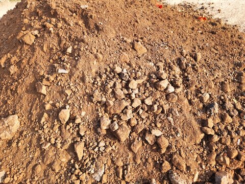Brown gravel and soil pile is situated on a construction site in an urban area, with scattered rocks and dust visible in daylight