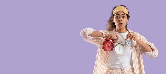 Overslept young African-American woman in pajamas holding alarm clocks on lilac background with...