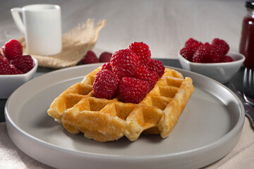 A waffle topped with raspberries on a gray plate, accompanied by bowls of raspberries, a white cup, and a jar in the background on a wooden table.
