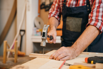 Carpenter in plaid shirt and blue overalls hammering a nail into a wood board on a workbench, surrounded by scattered tools and wood pieces, focused on precise craftsmanship, National Carpenters Day