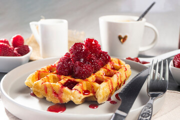 A golden waffle topped with fresh raspberries and raspberry sauce drizzling over it. The crispy...