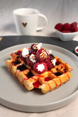 A waffle with raspberries, whipped cream, and raspberry sauce on a gray plate, accompanied by a cup of coffee, cutlery, and bowls with raspberries