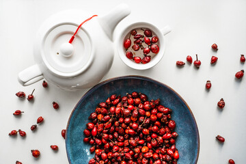 Rose hip herbal tea in a cup with a teapot and dried red berries on an around blue ceramic plate on a white wooden table. Medicinal plants, immune system support. Dog Rose in mug. Close-up top view.