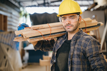Smiling construction worker in a yellow hardhat and plaid shirt holding wood planks on his...