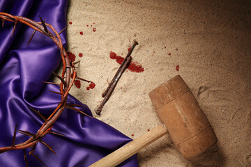 Crown of thorns with purple fabric, nails and hammer on sand background
