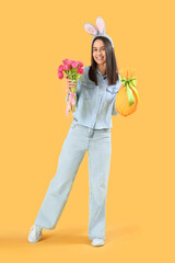 Young woman in bunny ears with tulips and Easter gift egg on yellow background