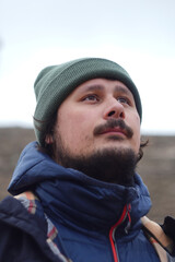 A contemplative man gazes upward, wearing a green beanie and a blue jacket. His expression reflects...