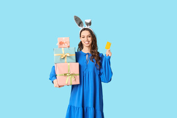 Happy young woman in Easter bunny ears headband with gift boxes and credit card on blue background