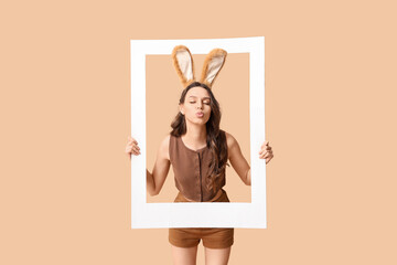 Young woman in Easter bunny ears headband with paper frame blowing kiss on beige background