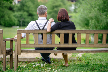 A middle-aged couple sits closely on a rustic wooden bench, surrounded by lush greenery, embracing...