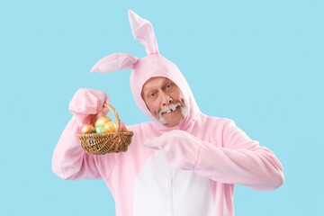 Senior man in Easter bunny costume pointing at wicker basket with painted eggs on blue background